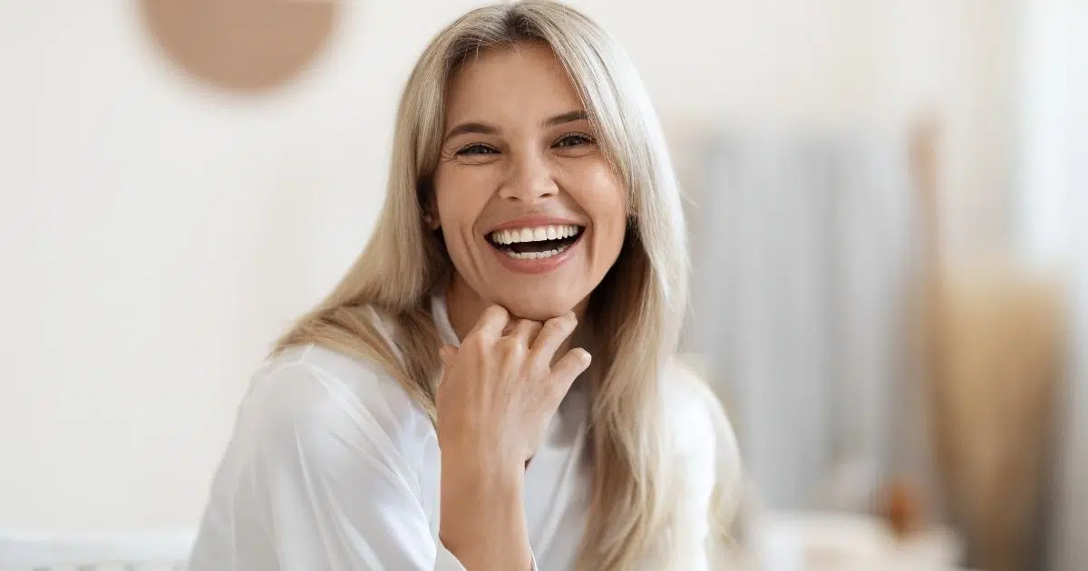Smiling woman with long blonde hair in a bright, comfortable indoor setting.