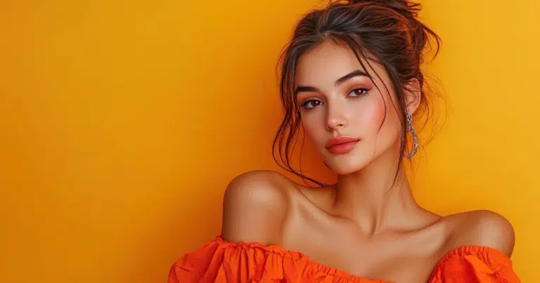 Portrait of a woman with a messy bun, dangling earrings, and an orange off-the-shoulder top against a vibrant yellow-orange background.