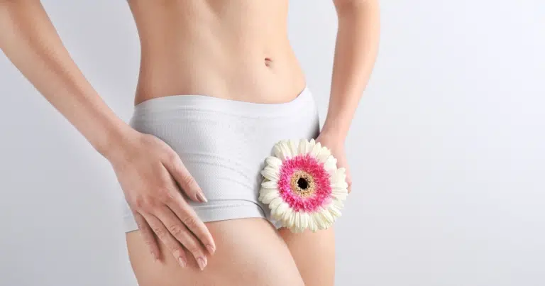Close-up of a woman's midsection wearing white underwear, holding a pink and white flower near the pelvic area, symbolizing feminine health and intimate wellness.